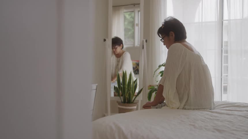 A woman with short dark hair and glasses wearing a light colored top sits on a bed looking towards a tall mirror that reflects her and a nearby potted plant in a bright room with a large window
