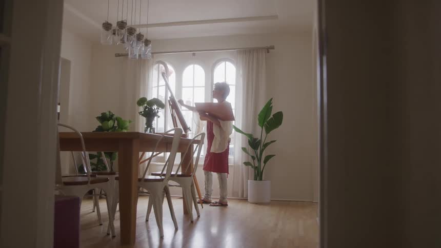 A woman paints on a canvas inside a dining room beside the window at midday