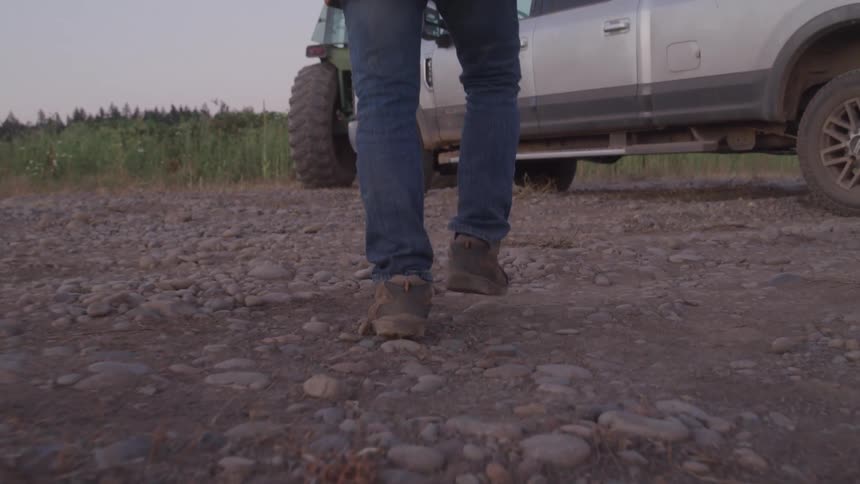 A person wearing jeans and work boots walks along a dirt road covered in small rocks with a white pickup truck and a tractor in the background showing large wheels