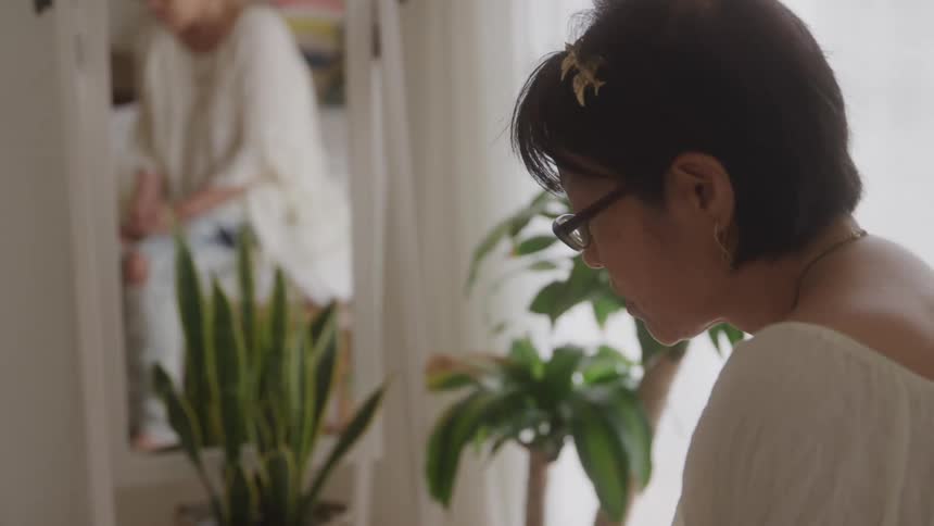 A woman wearing glasses and a white dress stands indoors in a room with a potted plant at midday