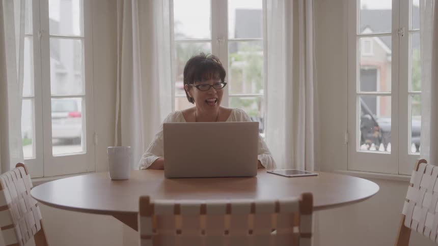 An asian woman smiles and laughs on a video call using a laptop while seated at a dining table indoors during daytime