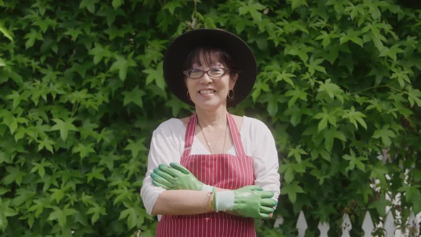 A woman wearing an apron hat and glasses stands in a garden at midday with a smile on her face in front of a tall plant with vibrant green leaves