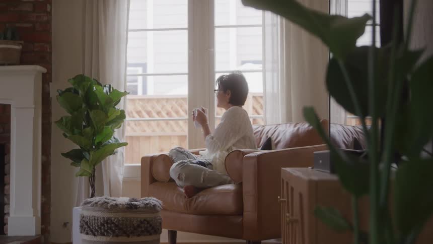 A woman sitting on the couch indoor holding a cup of coffee while looking through the window in the morning