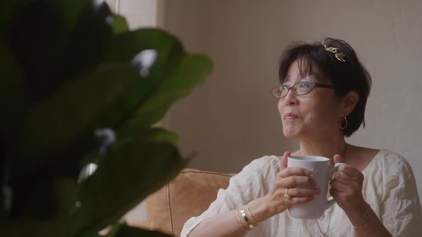 A woman with short dark hair and glasses wearing a light colored top and jewelry sits holding a white mug looking thoughtfully off to the side with green leaves of a plant in the foreground