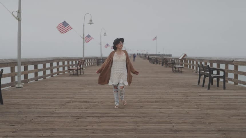 Woman in black hat putting her hands on her head walking along a wooden dock on a cloudy day