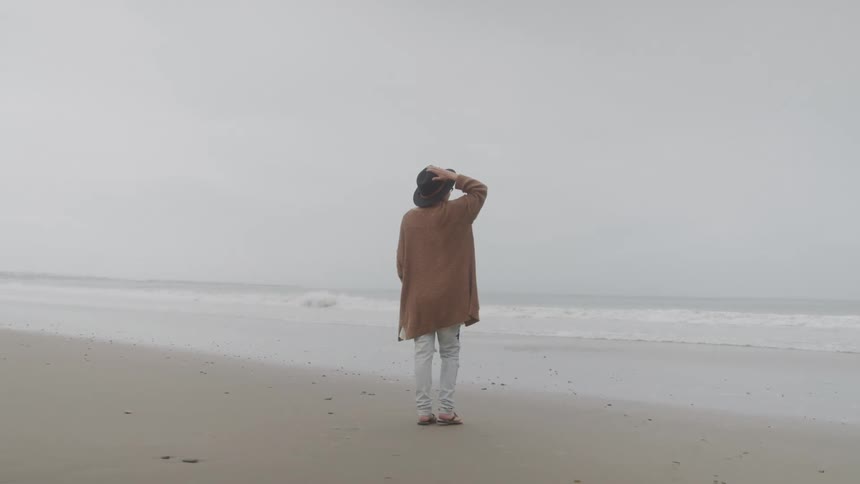 Person wearing a hat with his hand on his head walking on the sand towards the sea on a cloudy day