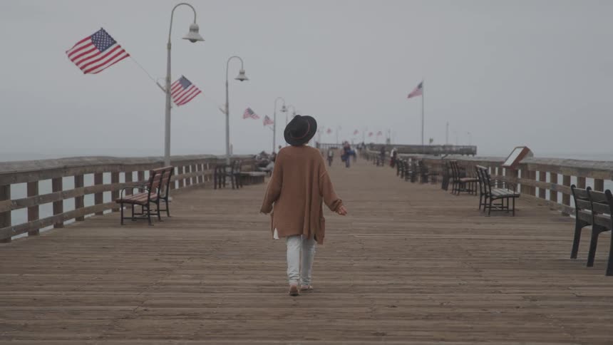 A person in a brown coat and hat walks away from the camera on a wooden pier adorned with american flags and street lamps the pier extends into a foggy or hazy expanse with benches lining both sides