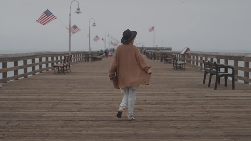 Person in a brown coat and black hat walks along a dock on a cloudy day