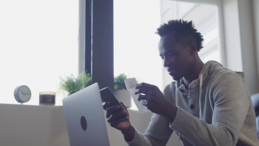 Man sits indoors holding a credit card and phone looking at screen