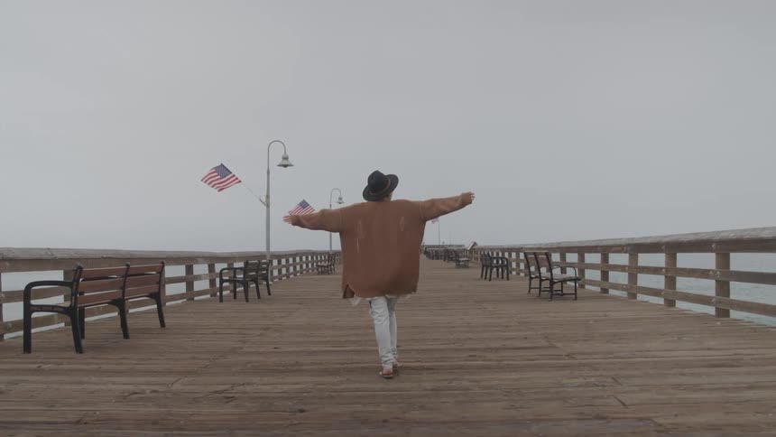 Person wearing a hat walking on a wooden dock on a cloudy day and opening his arms