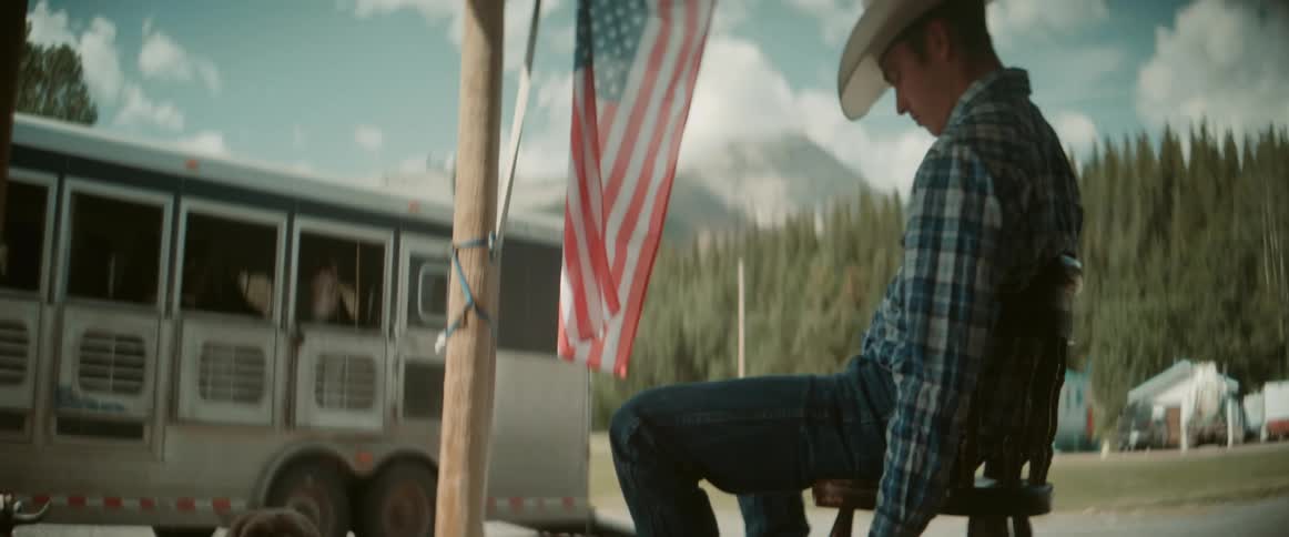 A brown dog lies calmly on a wooden porch next to a persons legs who is seated on a stool with a glass in hand in the background theres a white trailer with horse stalls and an american flag hanging from a pole