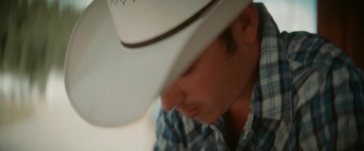 A man wearing a cowboy hat and a plaid shirt looks down with a pensive expression then sips from a clear glass and finally looks forward with a blurred outdoor background