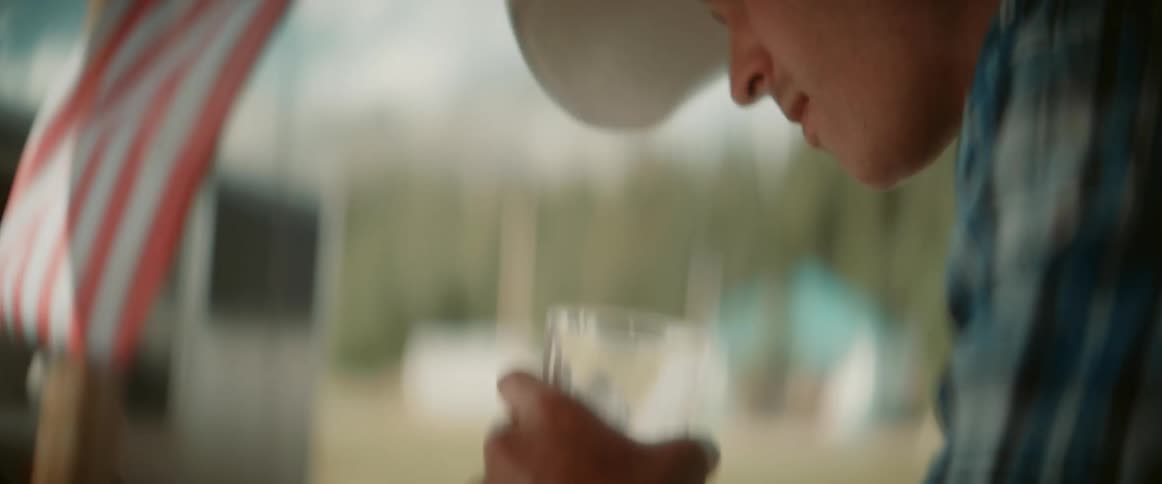 A man standing on the front porch of a building takes a sip from a glass of beer while wearing a cowboy hat with an american flag flying nearby at midday