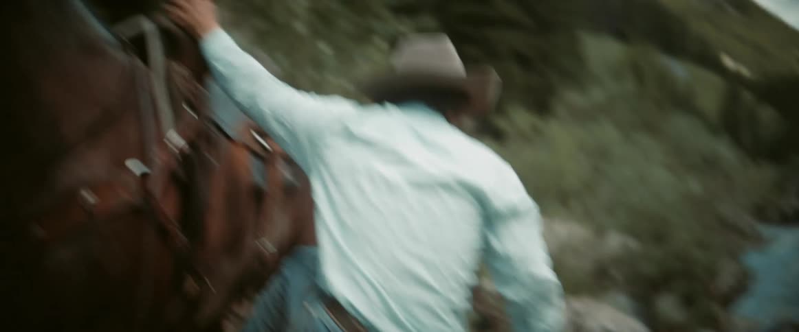 A cowboy wearing a lightcolored shirt and a hat is riding a horse looking forward with a serious expression the background features blurred green trees and a mountainous landscape