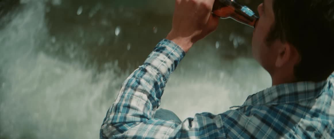 A person wearing a blue and white plaid shirt is shown from the shoulders up tilting their head back to drink from a glass bottle in the background water can be seen moving rapidly suggesting they are near a stream or waterfall