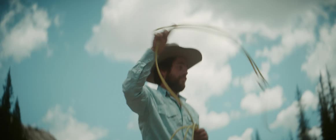 Cowboy twirls a lasso before attempting a throw while in a valley between conifer forest covered mountains on a cloudy day