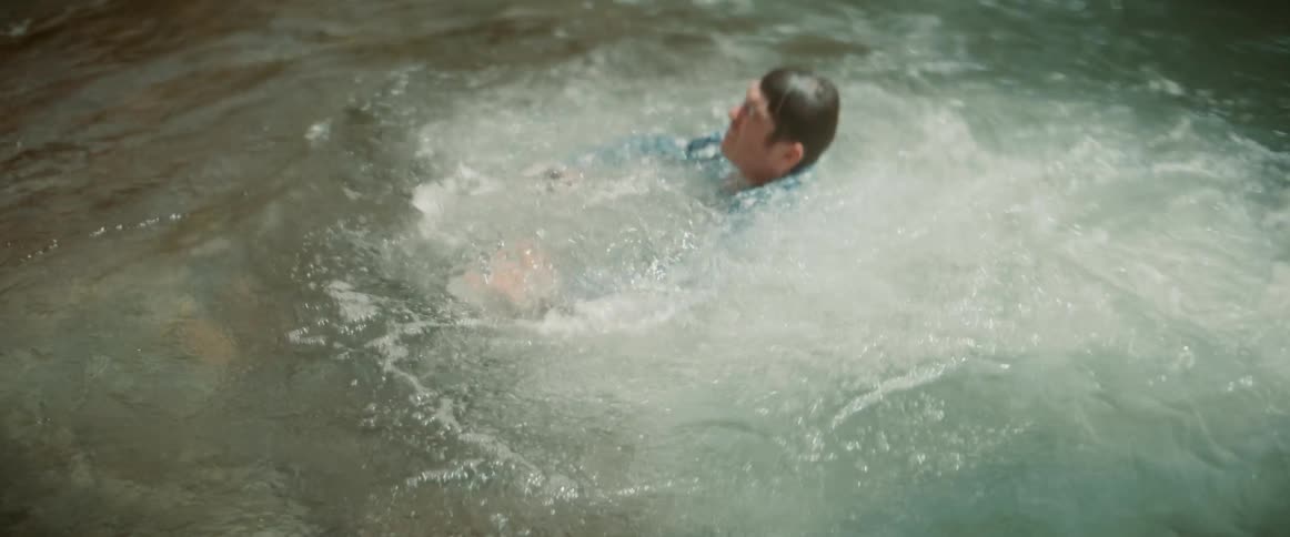 A person in a blue and white plaid shirt is seen from behind leaning forward and then standing upright facing a body of water that churns with white rapids the water is turbulent creating a dynamic background