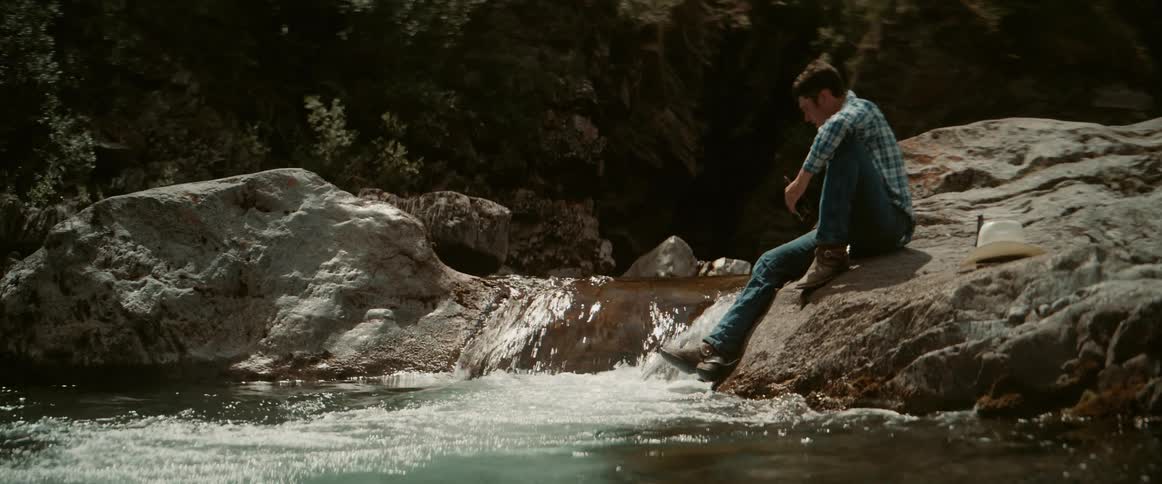 A man wearing jeans and boots sits on a rock beside a stream drinking a bottle of beer at midday