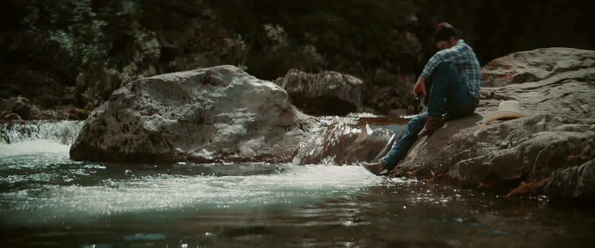 Intoxicated man sitting on the riverbank during the daytime falls into the water while holding a beer bottle
