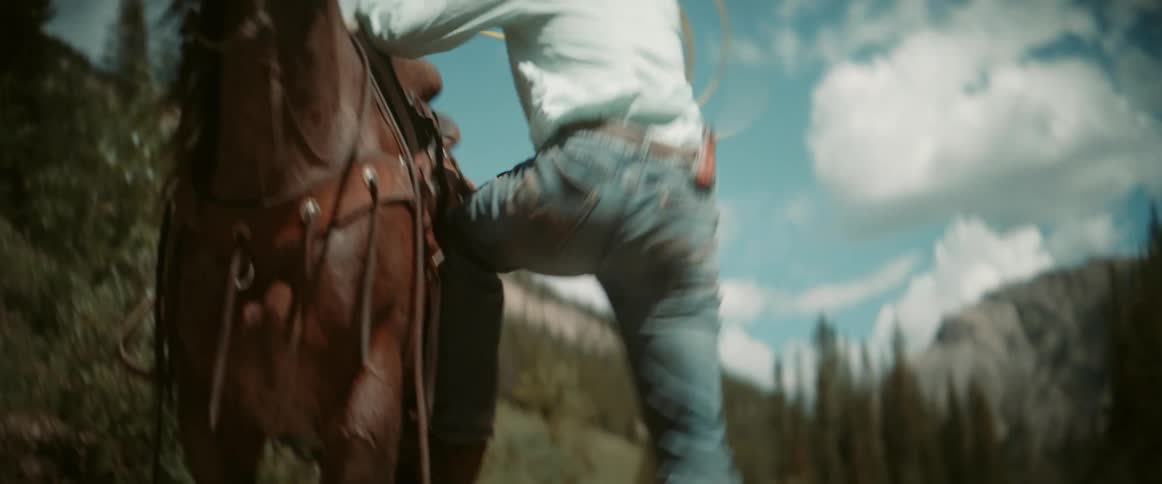 A man wearing a cowboy hat and boots climbs down from a horse he was riding holding a rope and walks toward a river