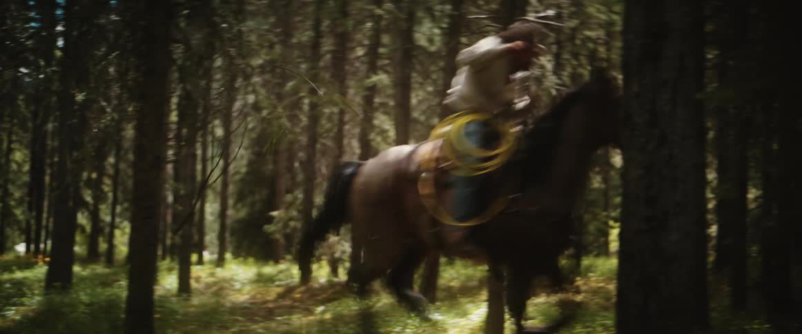 Two cowboys ride through a forest on horseback