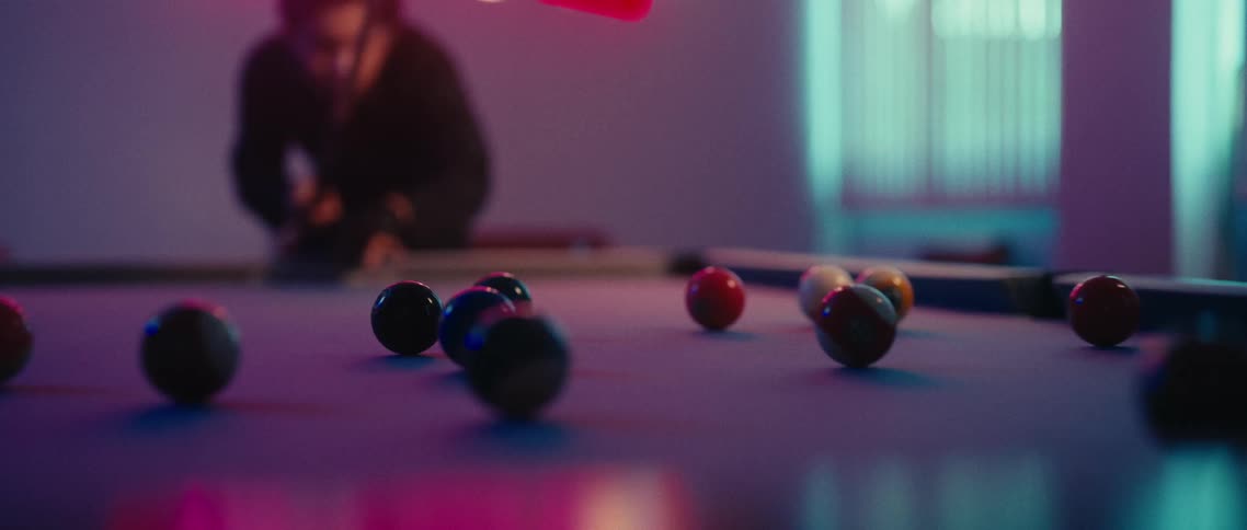 Male pool player makes a break shot while playing in a room lit by purple lighting