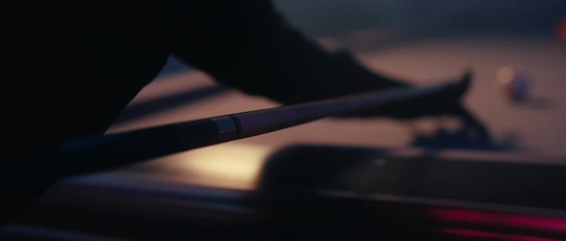 A person is holding and rotating a pool cue in their hands the cue reflecting various colored lights from the environment the background is dark and blurred suggesting a lowlight setting possibly a pool hall or bar