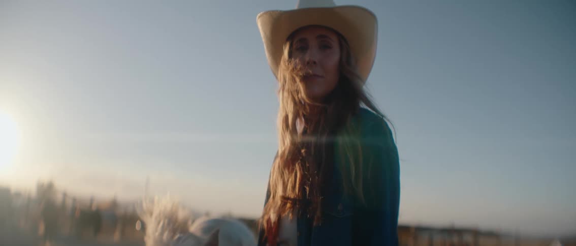 A young woman standing in an open area wearing a cowboy hat beside a white horse at sunset