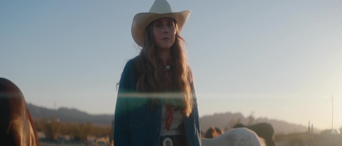 A person in a cowboy hat and blue jacket stands in a dusty open field next to a horse the background shows mountains under a clear sky with the sun low on the horizon