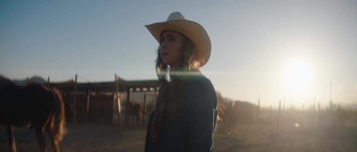 A woman with long hair wearing a cowboy hat and a dark shirt is seen in profile looking out at a hazy dusty ranch with horses and fences as the sun sets in the background