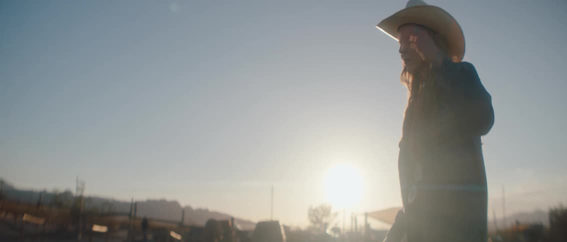 A woman wearing a cowboy hat and a dark jacket is seen in profile looking out at a bright hazy sunset with distant structures in a rural landscape