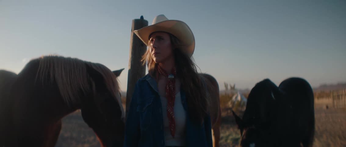 A person wearing a cowboy hat and a denim jacket stands between two horses looking towards the right side of the frame the background is blurred showing what appears to be a natural open landscape with a hint of a sunset or sunrise glow