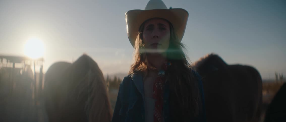 A cowgirl in a hat is shown in a closeup illuminated by the warm glow of the setting sun with blurred horses and a wooden post in the background her gaze is directed upwards creating a contemplative and serene mood