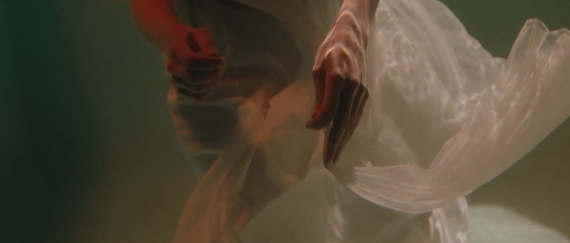A close view of a persons body is partially submerged in water with light colored fabric swirling around their arms and legs