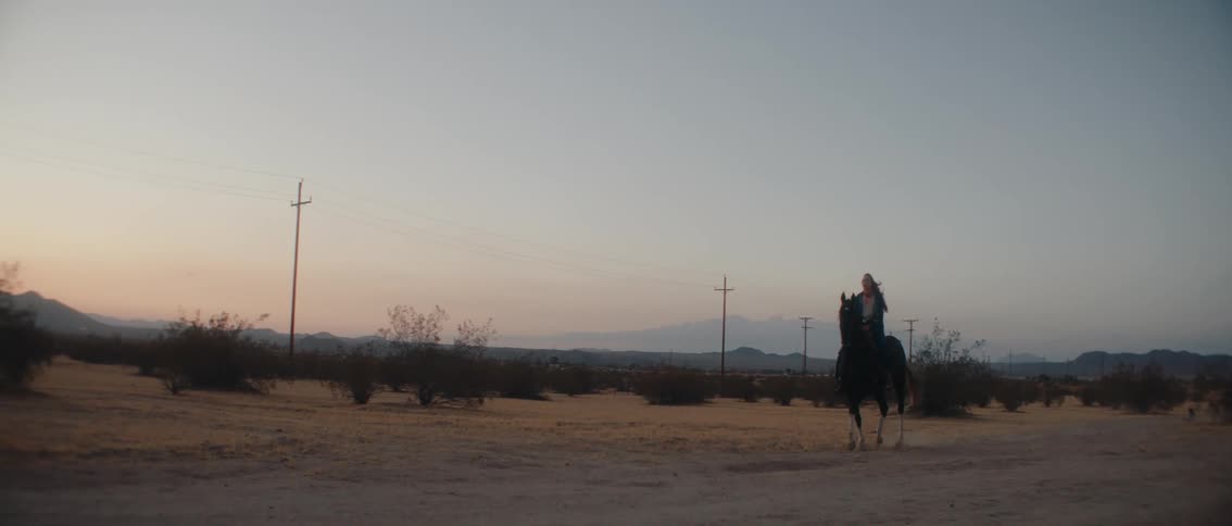 A lone woman on horseback slowly rides across a wide flat desert landscape under a hazy sunset or sunrise with power lines and distant mountains framing the serene solitary scene