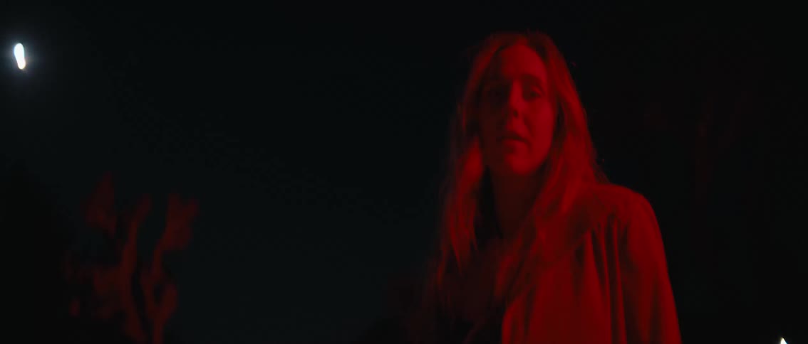 A lowangle shot of a mysterious womans feet and legs bathed in a deep red light transitions to a wider view revealing her face and a distant moon all while a sense of foreboding builds