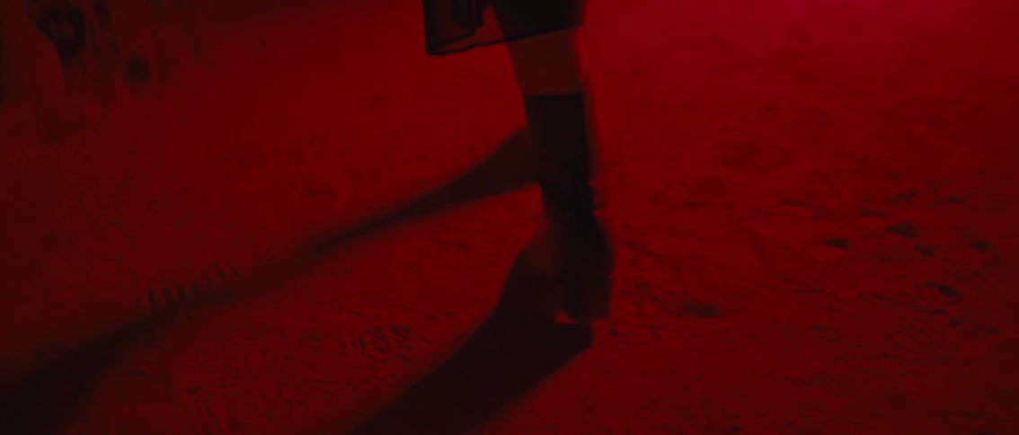 The video shows a pair of feet in dark possibly leather boots walking on a textured red surface casting a long shadow to the left the strong red hue of the ground and the clear visibility of individual textures create an intense and focused visual