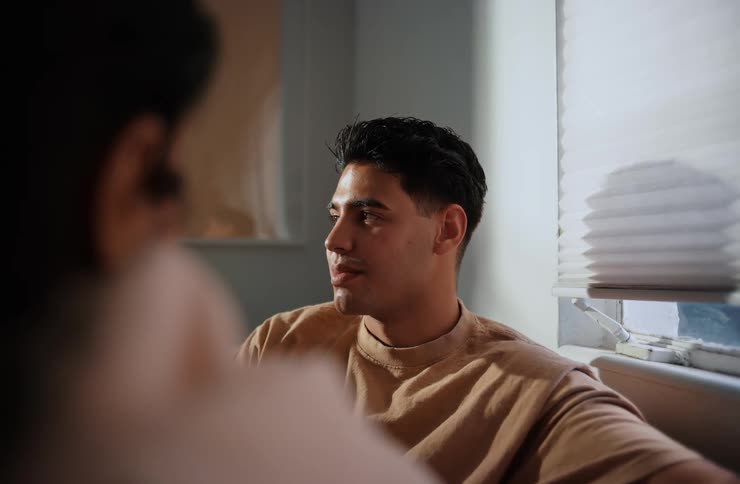 A man with dark hair wearing a light brown shirt sits and looks to his left seemingly conversing with another person whose blurred back is in the foreground with a window and blinds in the background