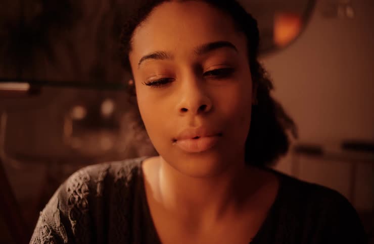 A young woman with a warm expression and her hair pulled back is looking directly at the camera in a closeup shot with soft indoor lighting