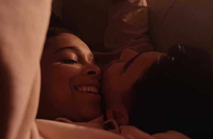 Two young adult women lie close together smiling and looking at each other in a dimly lit setting
