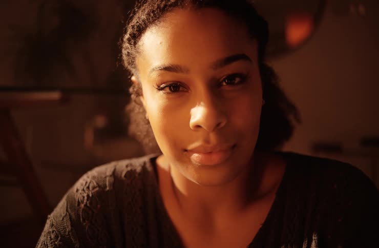 A young woman with a warm expression and her hair pulled back is looking directly at the camera in a closeup shot with soft indoor lighting