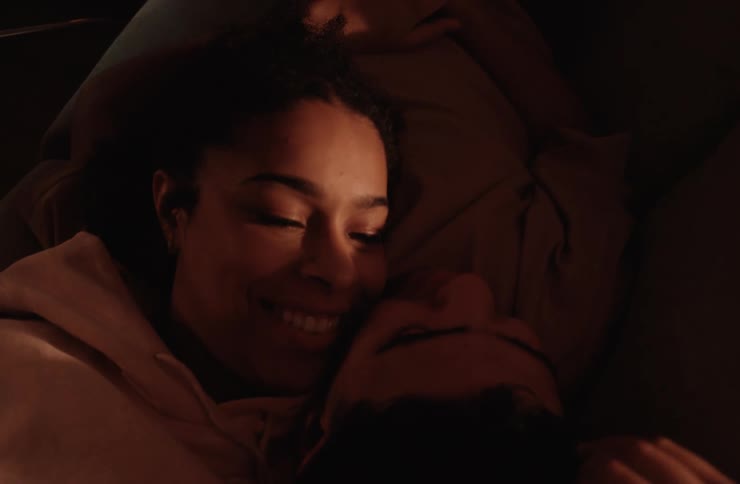 A young couple lie close together smiling and looking at each other in a dimly lit setting