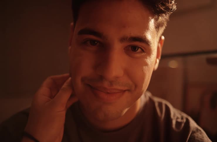A closeup shot of a man with a gentle smile with one hand resting on his cheek looking into the camera in a warmly lit indoor environment