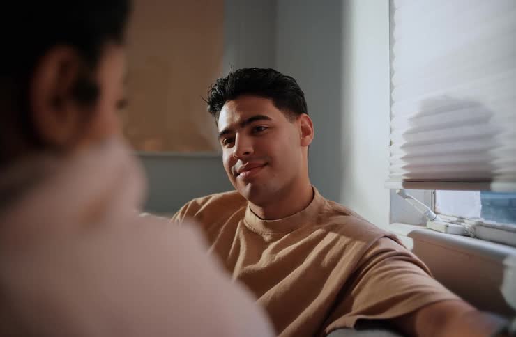 A shot of a smiling man in a tan shirt looking to his left with the blurry back of another person in the foreground and natural light coming from a window with blinds behind him