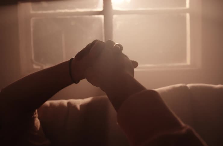 A silhouetted closeup shot of two peoples hands gently intertwined against a bright hazy and backlit window