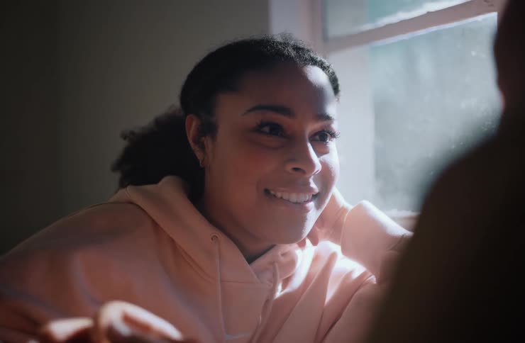 A young adult woman with dark hair in a ponytail wearing a light colored hoodie smiles broadly as she playfully holds hands with her boyfriend