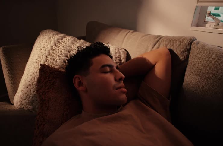 A medium shot of a man peacefully relaxing or sleeping on a couch with his head resting on pillows in a dimly lit room with a warm light source illuminating him from the side
