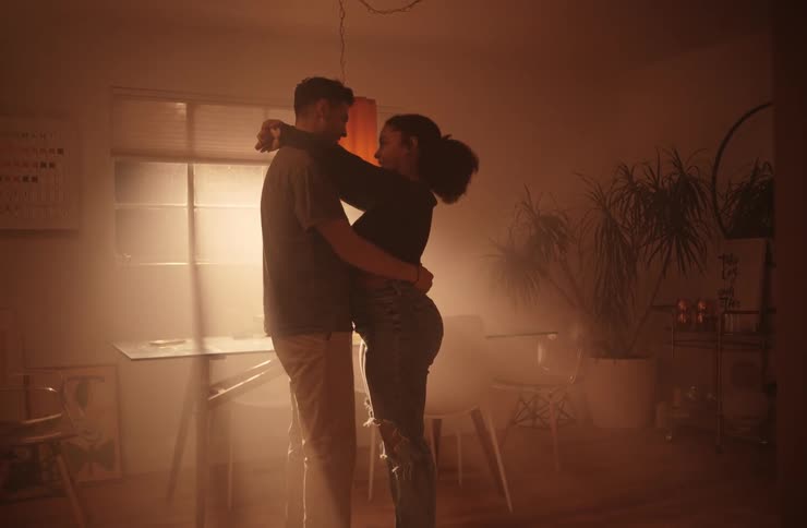 A couple is slow dancing in a dimly lit room holding each other and swaying gently they are looking into each others eyes and the room is filled with a hazy warm light