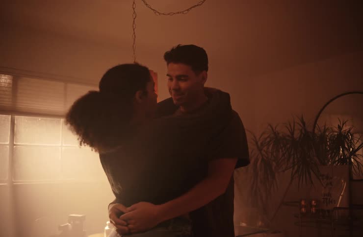 A couple is standing close together in a dimly lit room holding each other and looking into each others eyes they are smiling softly and appear to be sharing a tender intimate moment