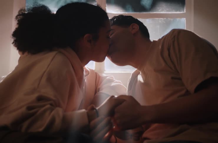 A young couple sitting on a couch beside a window at midday kissing and holding hands